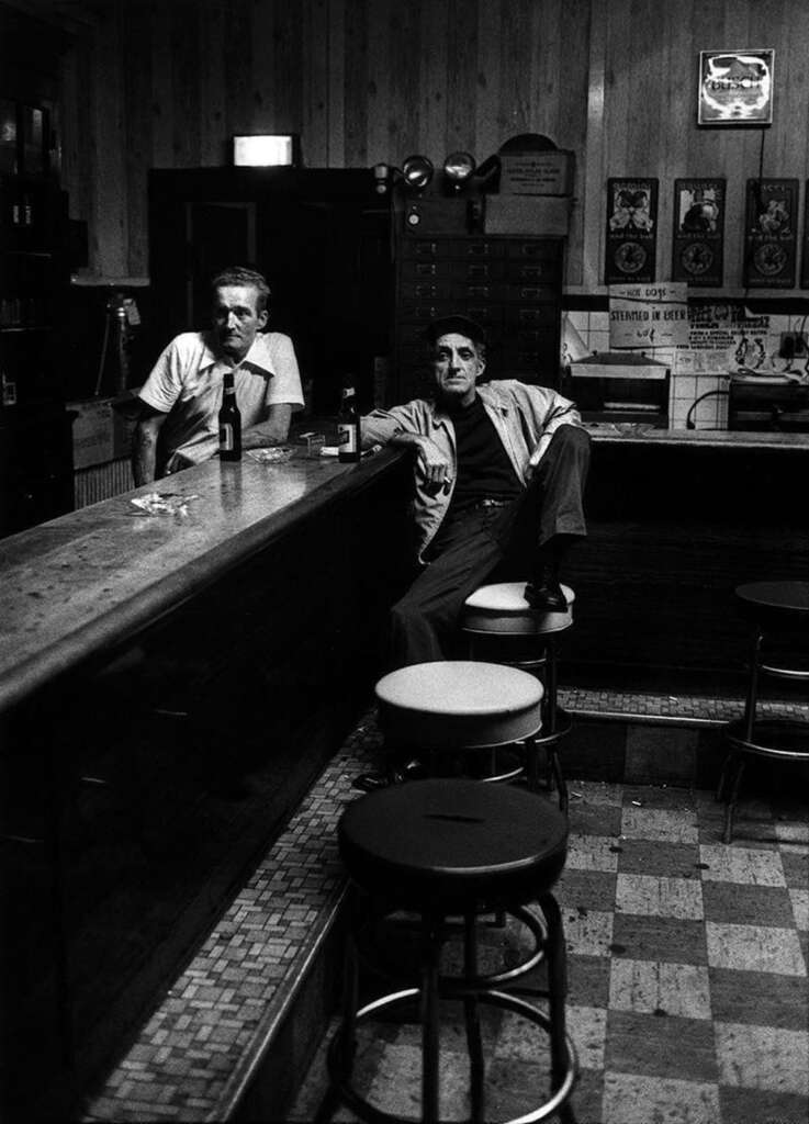 Two men in a dimly lit bar; one leans on the counter with a bottle, while the other sits on a barstool with a relaxed posture. The room features a wooden interior, framed posters on the wall, and a tiled floor.