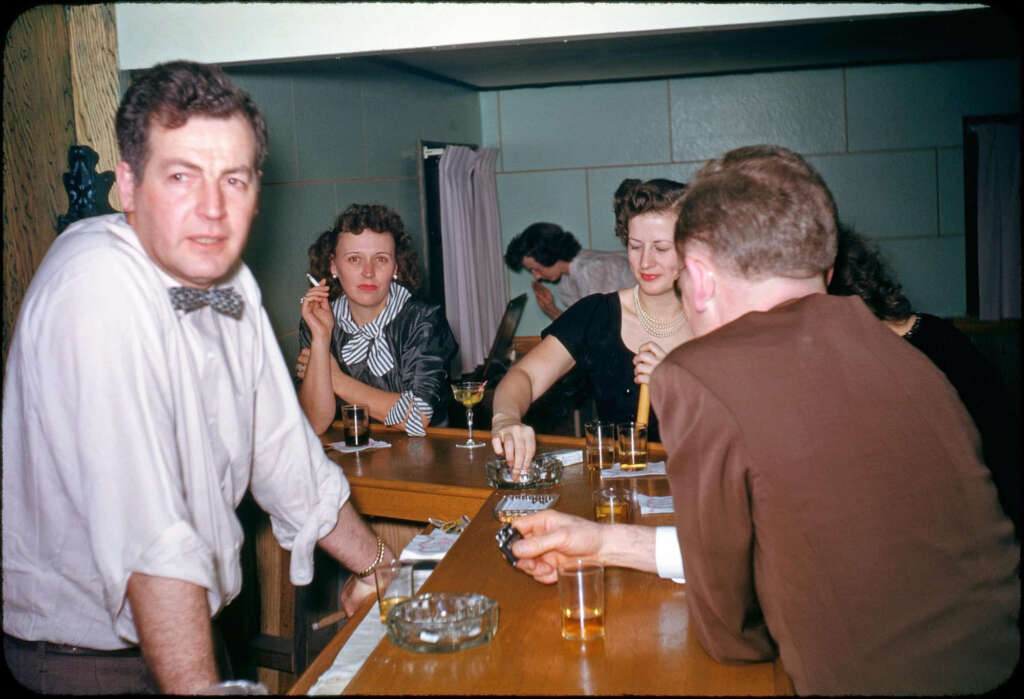 A group of people socializing at a bar. A man in a bow tie leans on the counter, while a woman smokes a cigarette. Glasses and ashtrays are on the bar. Others sit and chat in the background. The setting appears to be from the mid-20th century.