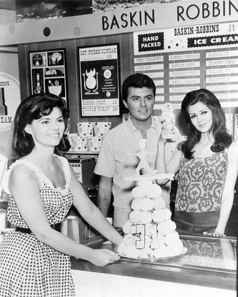 A vintage black-and-white photo of three people in a Baskin-Robbins store. Two women, one in a polka dot dress and another holding an ice cream cone, and a man in a collared shirt pose around a cake topped with a large "31" number.