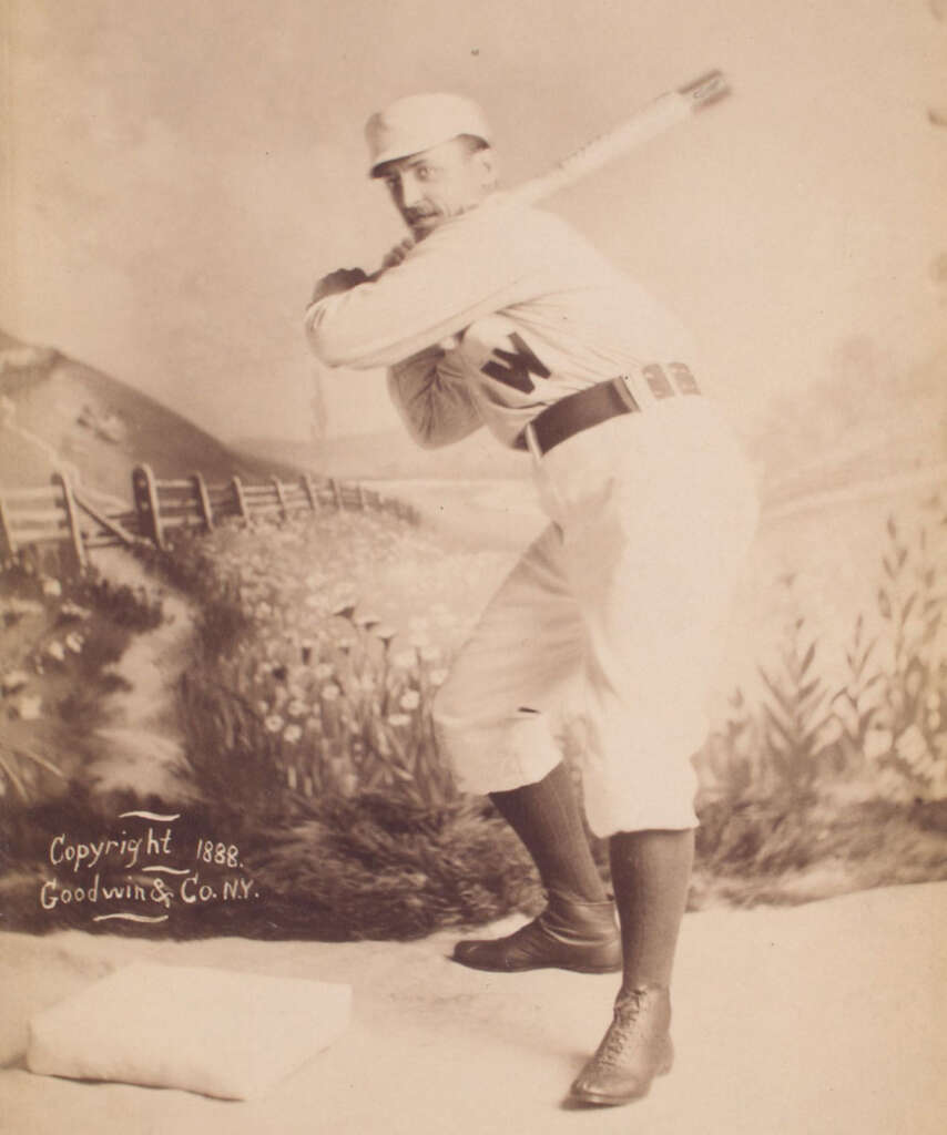 A vintage sepia-toned photograph from 1888 shows a baseball player in a batting stance. He's wearing a baseball uniform and cap, holding a bat over his shoulder. The background features a painted pastoral scene with a fence and flowers.