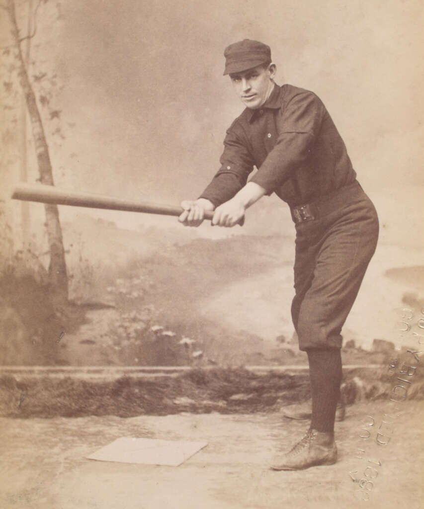 A vintage photograph of a baseball player in an early 20th-century uniform, posing with a bat as if about to swing. He is wearing a cap and standing near a base on a field with a blurred natural backdrop.