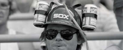 A person wears a helmet with "SOX" on it, holding two cans connected by straws for drinking. They are wearing sunglasses and sitting in a crowd. The image is in black and white.