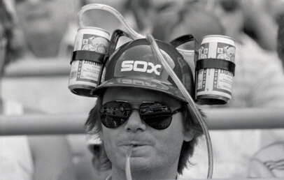 A person wears a helmet with "SOX" on it, holding two cans connected by straws for drinking. They are wearing sunglasses and sitting in a crowd. The image is in black and white.