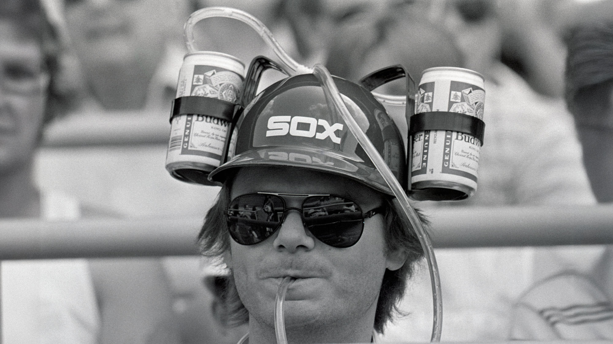 A person wears a helmet with "SOX" on it, holding two cans connected by straws for drinking. They are wearing sunglasses and sitting in a crowd. The image is in black and white.