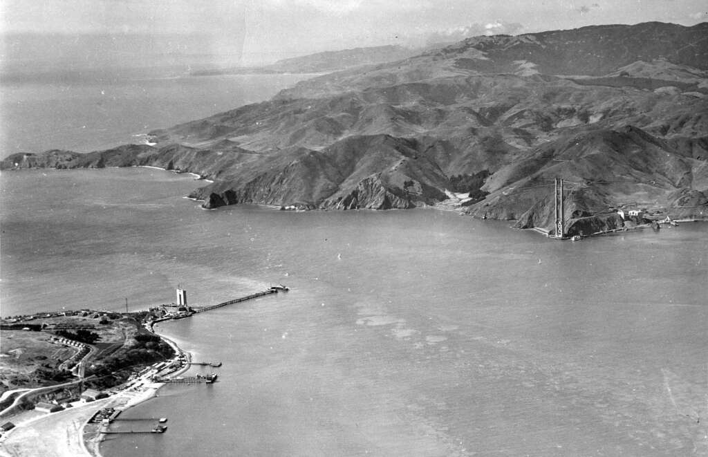 Aerial view of the Golden Gate Strait, showing the early construction of the Golden Gate Bridge with towers partially built. Surrounding hills and coastline are visible under a cloudy sky.