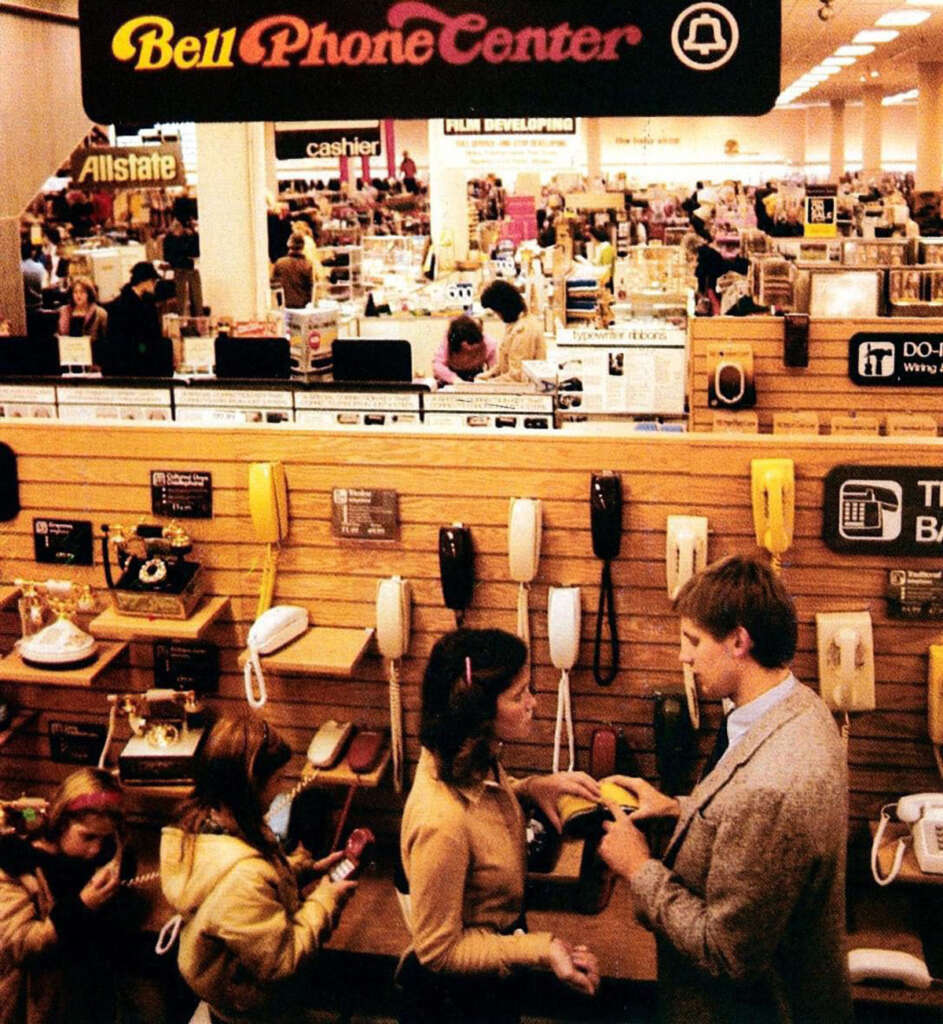 A vintage photo of a Bell Phone Center inside a busy department store. Various corded phones are displayed on shelves. A man and a woman are interacting at the counter. Shoppers browse in the background. Signs for Allstate and film developing are visible.