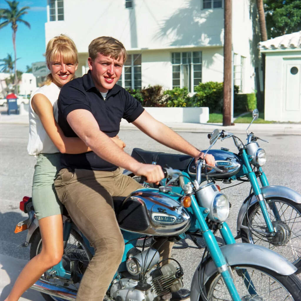 A man and woman sitting together on a blue vintage motorcycle in a sunny, suburban street. The woman smiles, holding onto the man. Another identical motorcycle is parked nearby. White houses and palm trees are in the background.