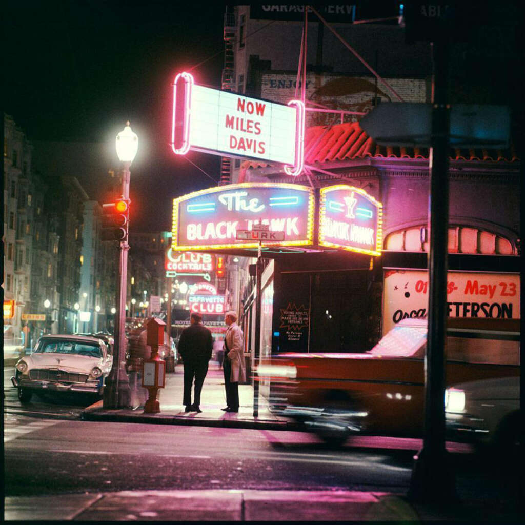 Nighttime street scene with neon signs and lights outside a jazz club called "The Black Hawk." A sign advertises "Now Miles Davis." Classic cars and two people on the street are visible, adding a vintage feel to the urban setting.