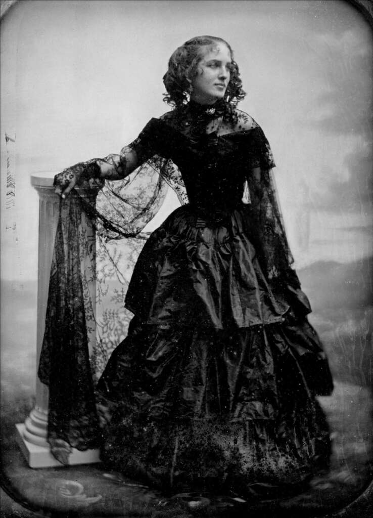 Black and white portrait of a woman in a Victorian-era gown, standing with one arm resting on a pedestal. She wears a long, dark, layered dress with lace details and a high collar, her hair styled in curls.