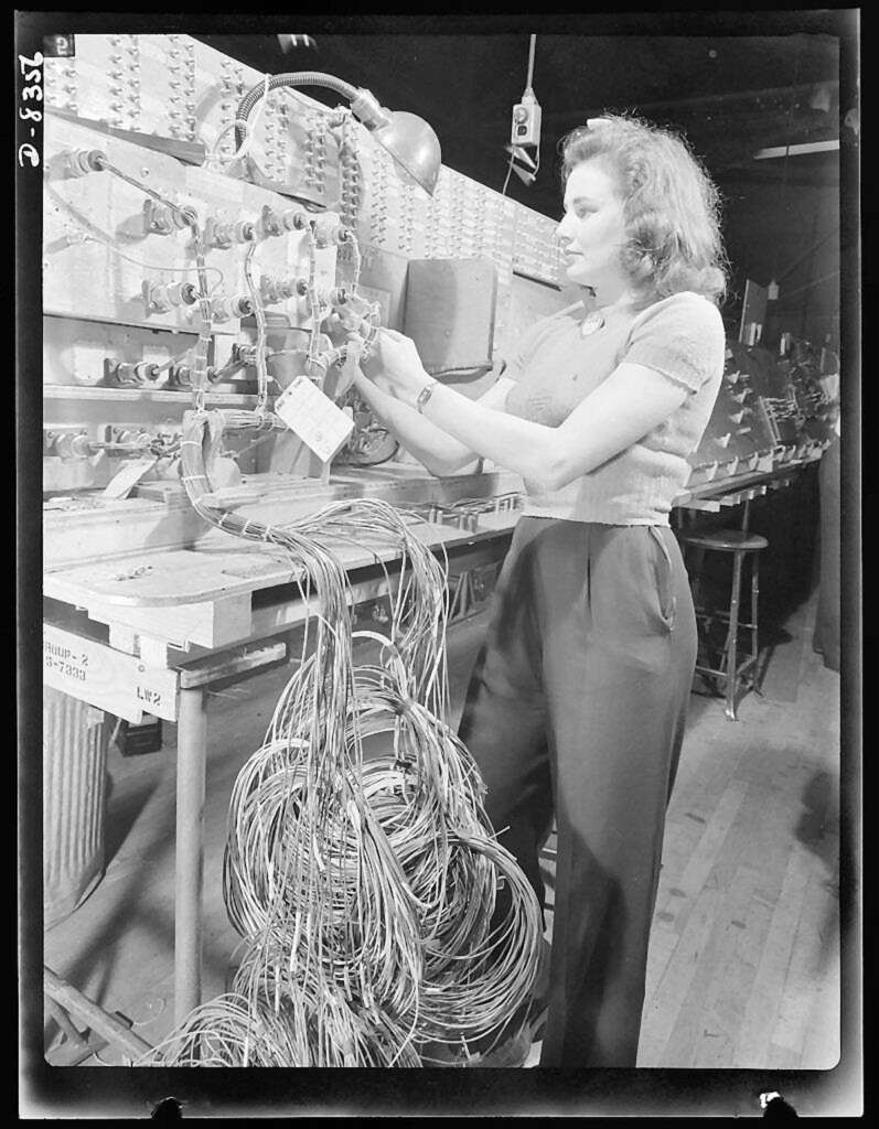 A woman operates an early computer, standing in front of a large panel with numerous wires and switches. She is adjusting connections, focused on her task. A tangled bundle of cables lies on the floor beside her. The setting has a vintage, industrial feel.