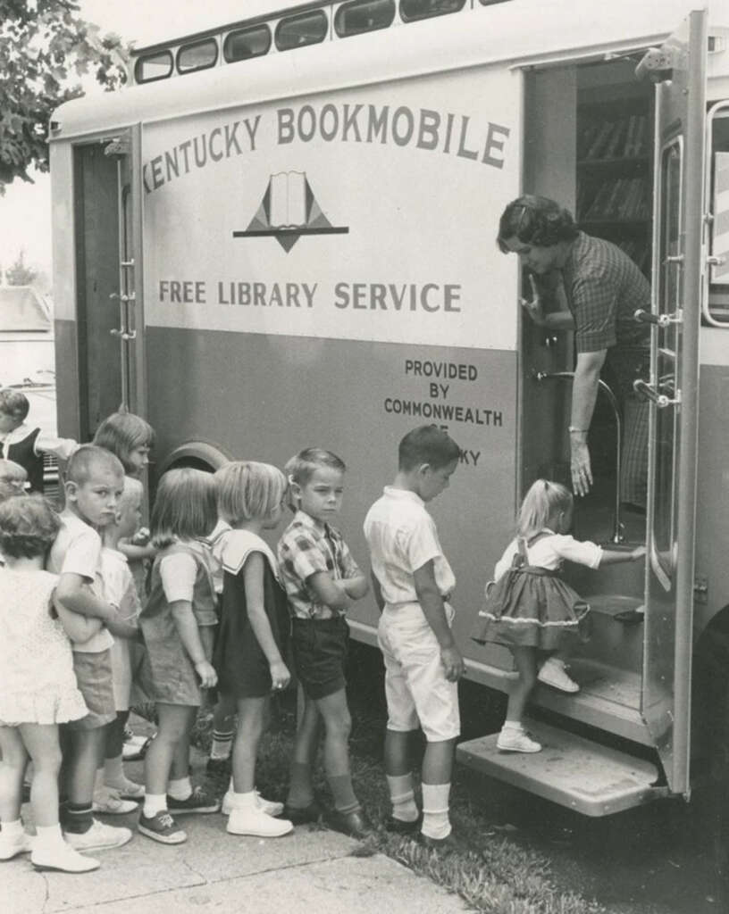 Children line up to enter a vintage Kentucky Bookmobile offering free library service. A woman assists a child stepping into the vehicle. The side of the bookmobile displays the service details, marked as provided by the Commonwealth of Kentucky.