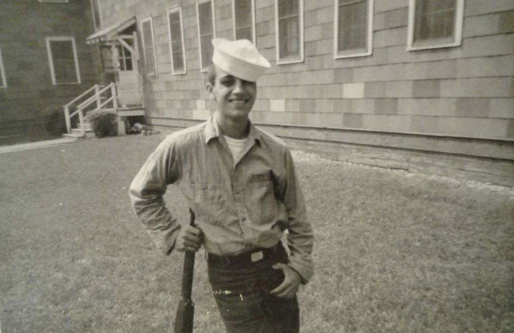 Young man in a sailor uniform stands smiling in front of a wood-paneled building, holding a rifle by his side. The image is black and white, suggesting a vintage feel. The man wears a cap tilted to the side.