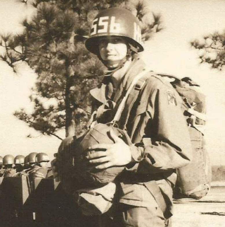 A sepia-toned image of a soldier wearing a helmet with "656" on it. The soldier is in uniform, holding a parachute pack, with trees visible in the background, suggesting a military training or paratrooper setting.