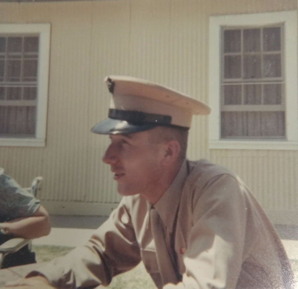 A man in a military uniform with a peaked cap is sitting outside next to a person in a floral shirt. A wooden building with two windows is in the background. The scene appears to be in daylight.