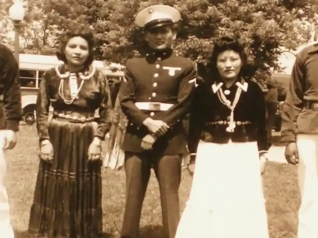 Three people stand outdoors. A man in a military uniform is flanked by two women in traditional dresses. They stand on grass, with trees and a vintage vehicle in the background. The photo has a sepia tone, giving it a historical feel.