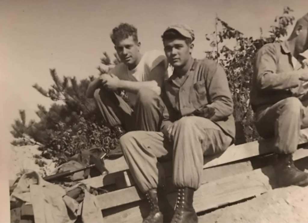 Two men in military attire sit outdoors on wooden planks. One has his arms resting on his knees, while the other wears a cap. Trees and bushes are visible in the background. Another person is partially visible on the right. The image is in sepia tone.