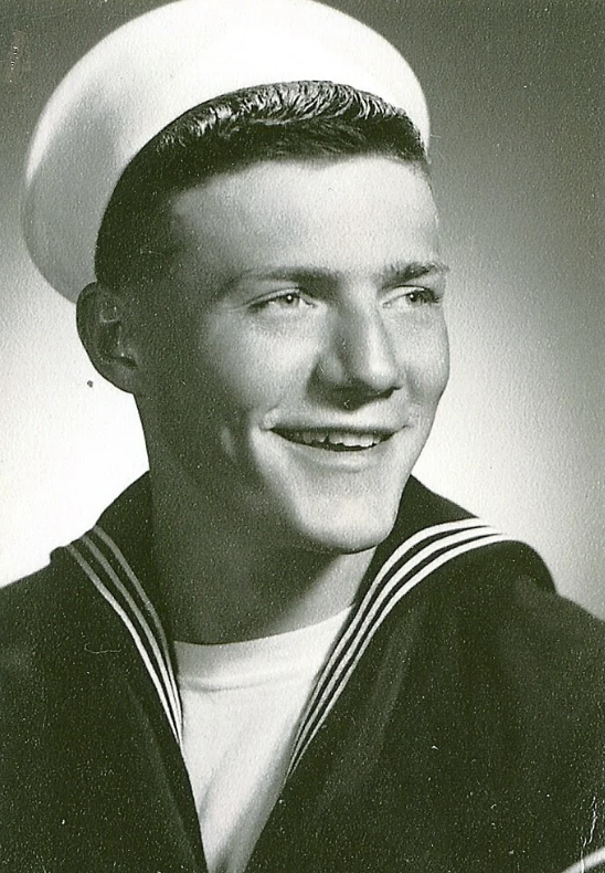 A young man wearing a sailor uniform, including a white hat and a dark jacket with light stripes on the collar, smiles while looking off to the side in a black and white portrait.