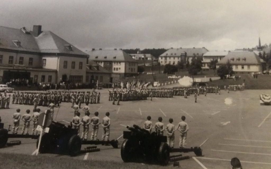 A black and white photo of a military parade, featuring soldiers lined up in formation on a large paved area. Several cannons are in the foreground, with buildings and trees in the background under a cloudy sky.