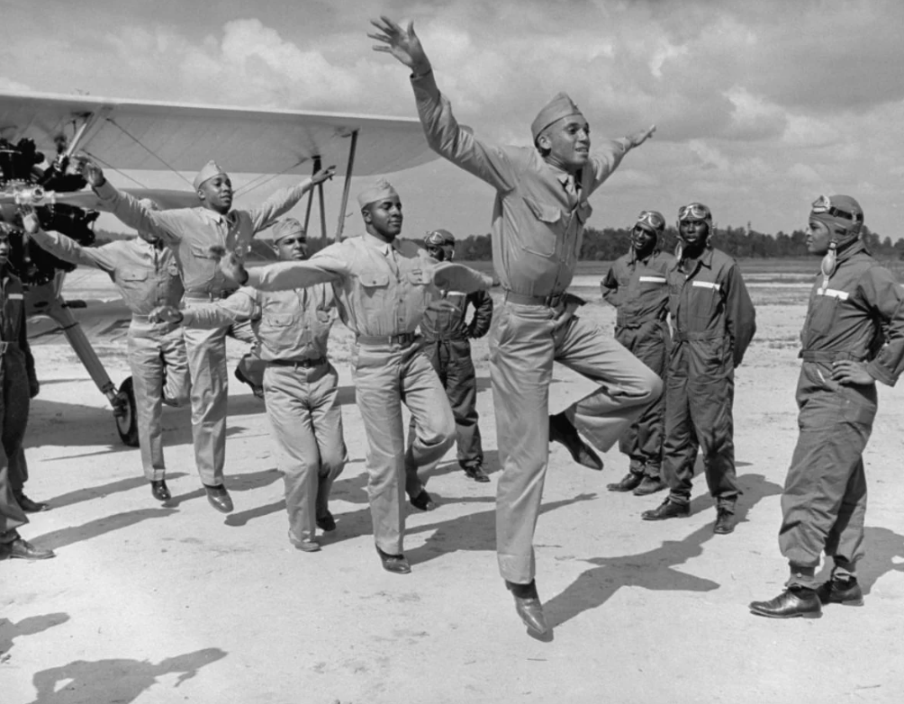 A group of pilots in uniform perform animated jumps in front of a biplane under a partly cloudy sky. Some stand observing, while others are mid-air with arms and legs extended. A line of trees is visible in the background.