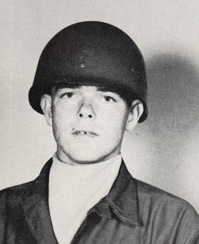 A young man in a military uniform and helmet poses for a portrait. He wears a high-collared shirt under his jacket. The image is black and white, giving it a vintage appearance.