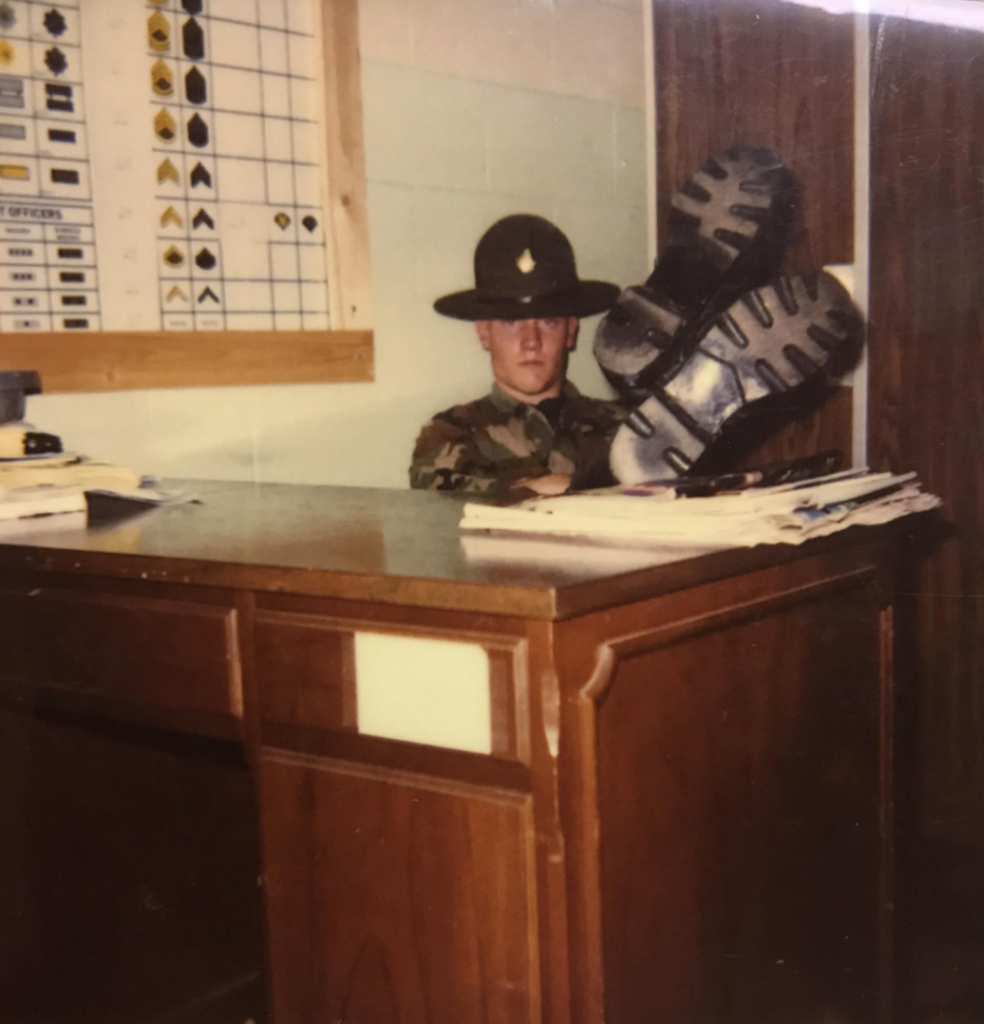 A person in a military uniform sits behind a wooden desk with one boot resting on the surface. A chart with military ranks is on the wall in the background. Papers and documents are scattered across the desk.