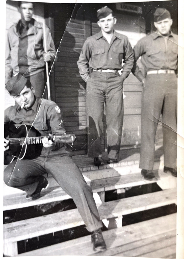 Four men in military uniforms stand on wooden steps outside a building. One man sits in front, playing a guitar, while the others stand behind him with relaxed postures. The image is black and white, with a vintage feel.