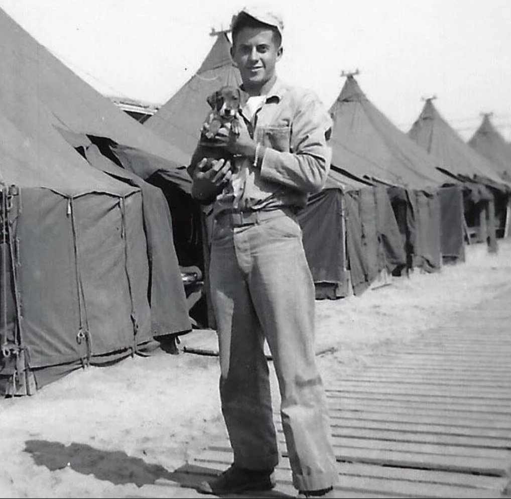 A man holding a small dog stands on a wooden walkway near a row of military tents. He wears a rolled-up shirt and jeans, with a cap on his head. The scene appears to be in a military camp.