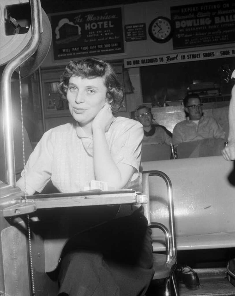 A woman sits at a counter, resting her chin on her hand, with a retro-style pin-setting machine nearby. Behind her, people are seated on chairs, and vintage signage for a hotel and bowling alley decorates the wall.