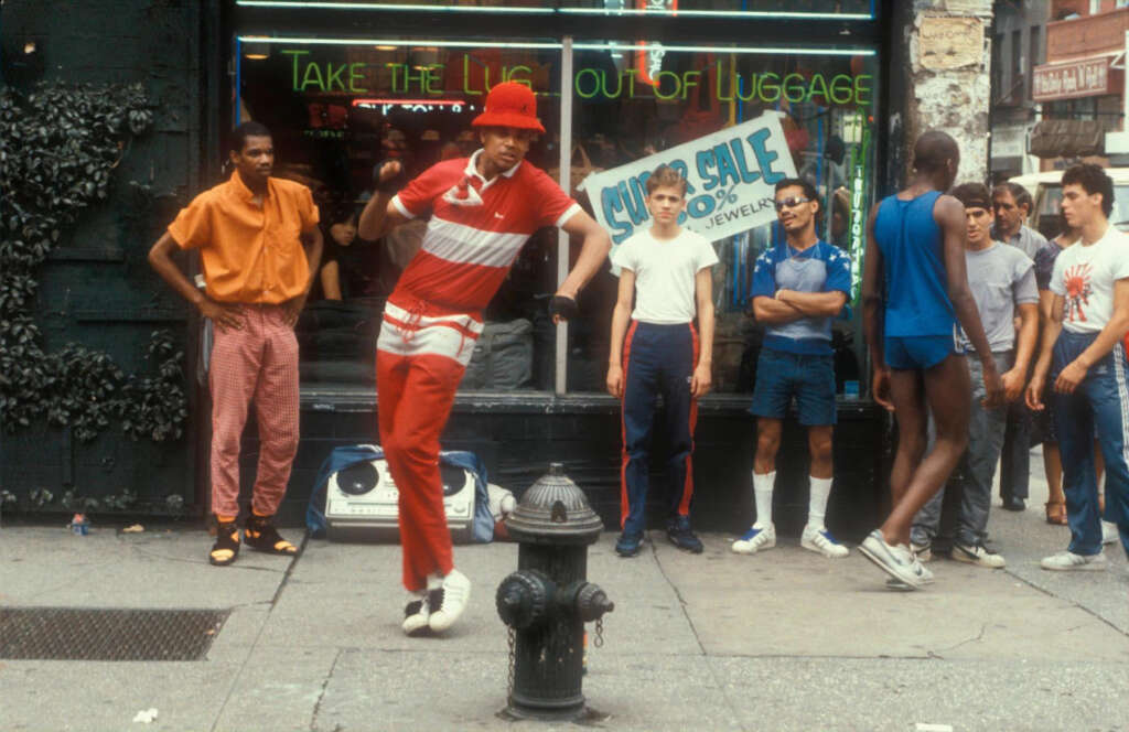 A person in a red and white outfit dances energetically on a city sidewalk, surrounded by a group of casually dressed spectators. A storefront behind them displays a sign about a sale on jewelry. A boombox is on the ground.