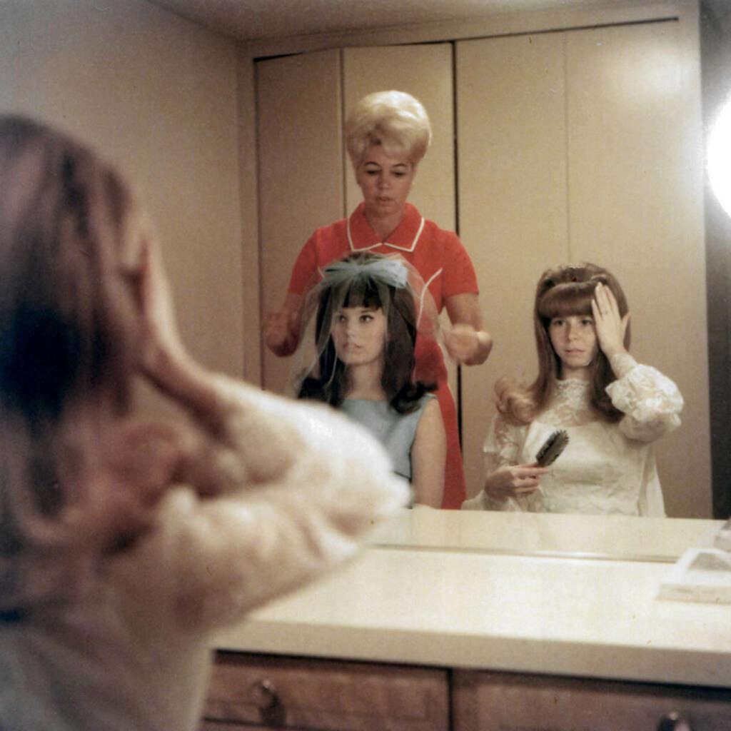 A woman in a red dress styles the hair of a seated woman with a blue ribbon, while another woman with a brush adjusts her hair. They are in front of a large mirror in a dimly lit room. A third person is blurred in the foreground.