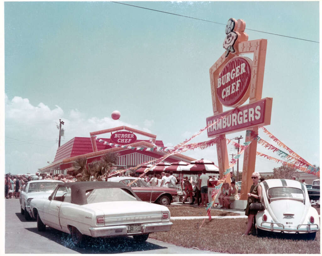 A vintage scene outside a Burger Chef restaurant with a large, colorful sign. Several people are gathered, and classic cars are parked, including a convertible and a vintage VW Beetle. Festive streamers add to the lively atmosphere.