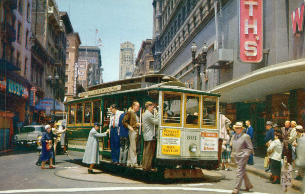 Streetcar in a bustling city intersection with people boarding and disembarking. Surrounding buildings display vintage signage. A car waits behind the streetcar, and pedestrians walk nearby. The scene captures a lively urban atmosphere.