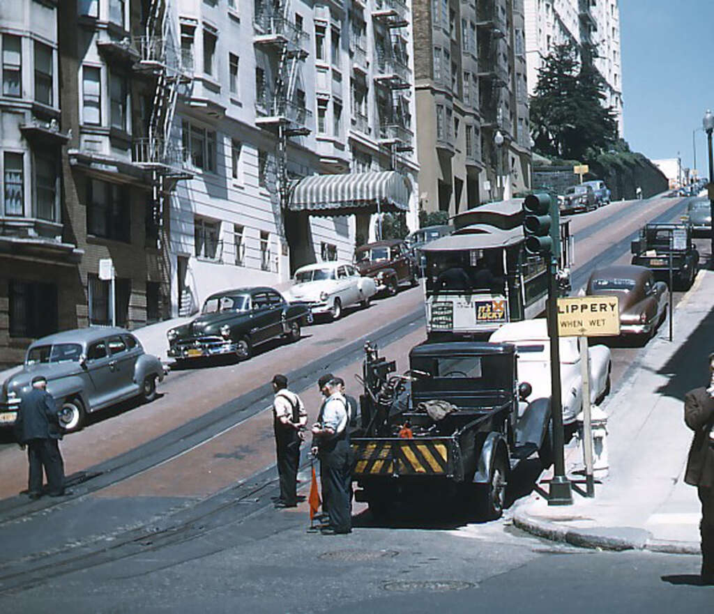 A steep city street with vintage 1950s cars parked along the side. An old cable car sits on the tracks, with two workers nearby. A "slippery when wet" sign is visible. Tall buildings line the street.