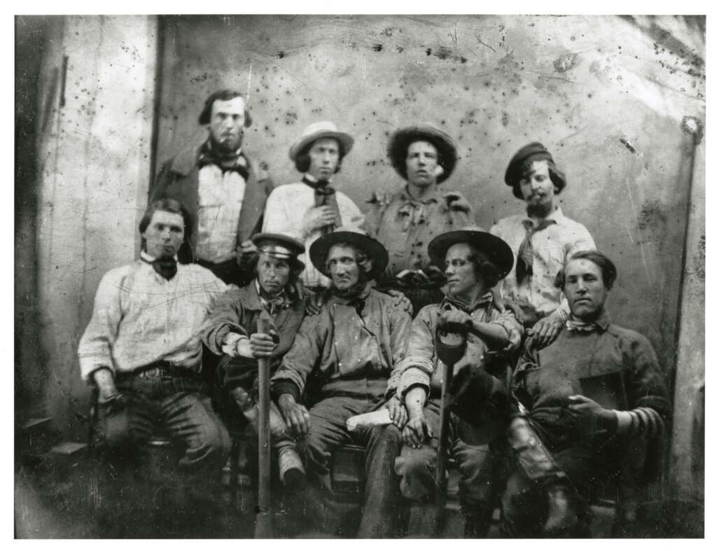 A vintage black and white photo of nine men in 19th-century attire, with diverse hats and clothing. Some sit holding rifles, while others stand behind them. The background is plain, with scratches and marks on the image, typical of old photographs.