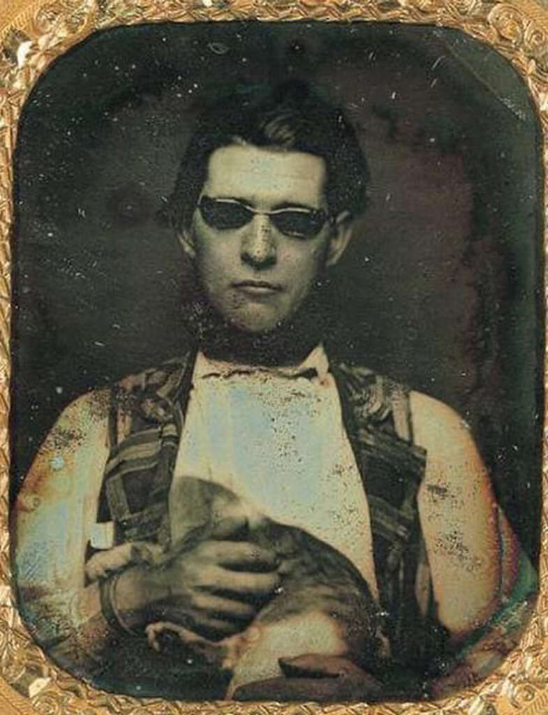 A vintage photograph features a person wearing dark sunglasses and a patterned vest over a shirt, holding a cat in their arms. The image has an old, sepia-toned appearance with a decorative frame around it.