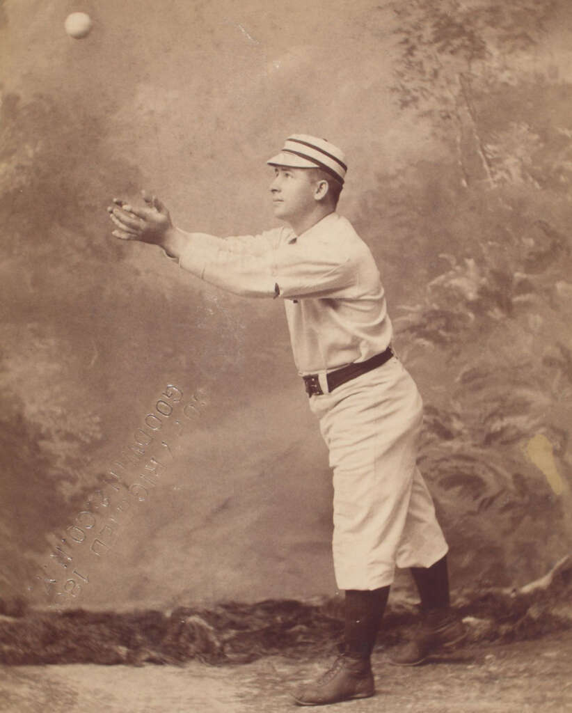 A vintage photograph of a baseball player wearing a striped cap and a uniform, poised to catch a ball in mid-air. The backdrop features a painted nature scene, giving a studio-like appearance.