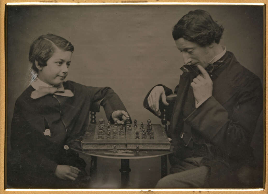 A young boy and an adult male are playing chess at a small round table. The boy appears to be making a move, while the man is observing intently, resting his chin on his hand. Both are dressed in formal attire. The image is in sepia tones.