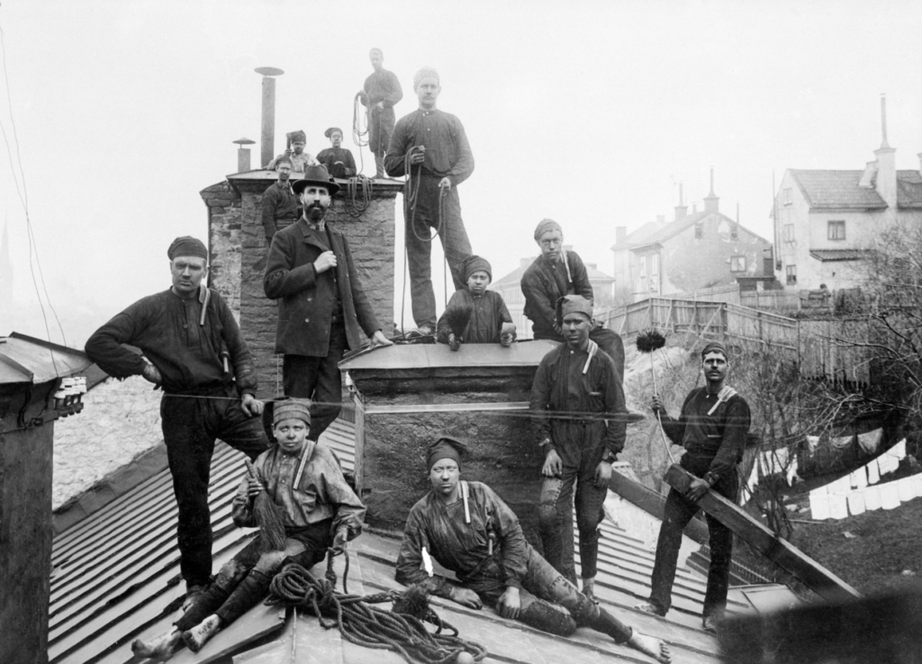 Historical black and white photograph of chimney sweeps on a rooftop. Eleven men and boys dressed in work clothes, caps, and holding tools, pose around chimneys. Houses and a clothesline with hanging laundry are visible in the background.