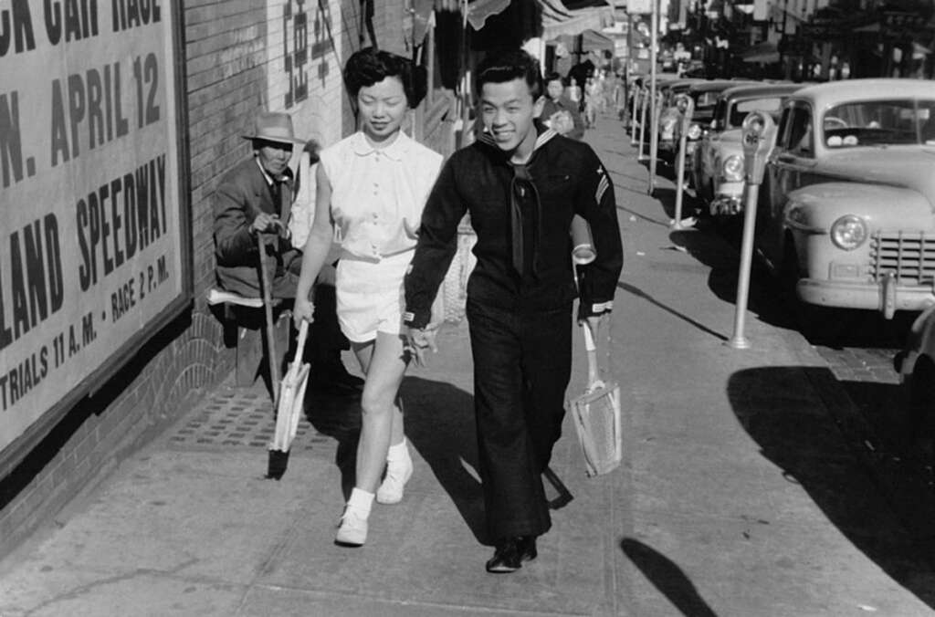 A smiling couple walks hand in hand on a city sidewalk. The man, in a black uniform, holds a newspaper. The woman wears a light-colored dress. Vintage cars and a street vendor are visible in the background.