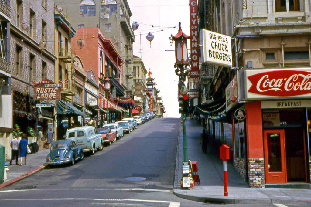 A vibrant city street with a steep incline is lined with vintage cars. Signs for businesses, including "Rustic Lodge," "Big Ben's Chuck Burgers," and "Coca-Cola," are visible. The architecture is a blend of classic and early modern styles.