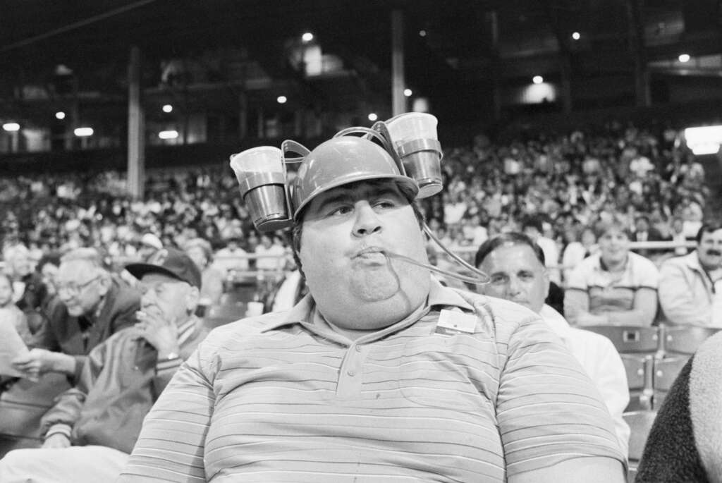 A man in a stadium wears a helmet with two beer cans attached and straws reaching his mouth. He's seated among a large crowd, and the image is in black and white.