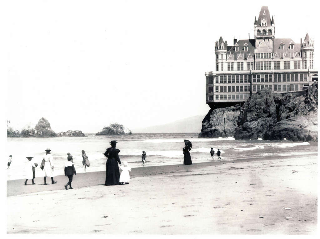 A vintage black and white photograph of people in old-fashioned attire walking along a beach. In the background, a large, ornate building perched on a rocky cliff overlooks the ocean waves.