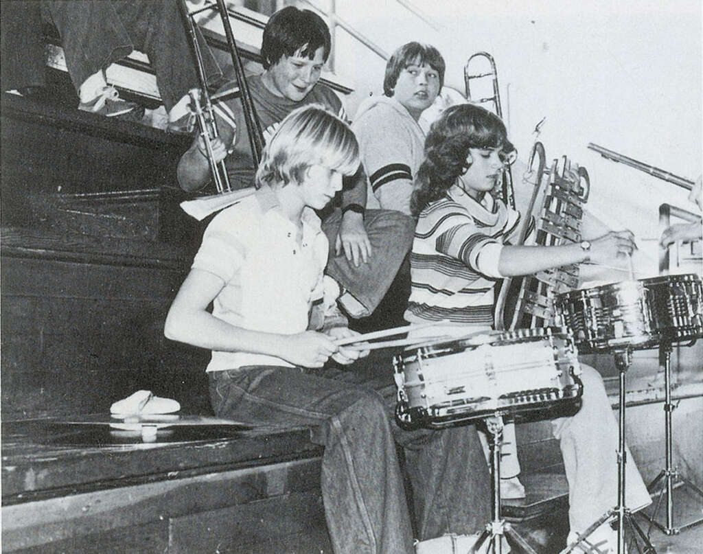 A group of young people sits on bleachers with musical instruments. One plays the drums, while others hold a trombone and cello. They're in a casual setting, possibly at a school. The image is in black and white.