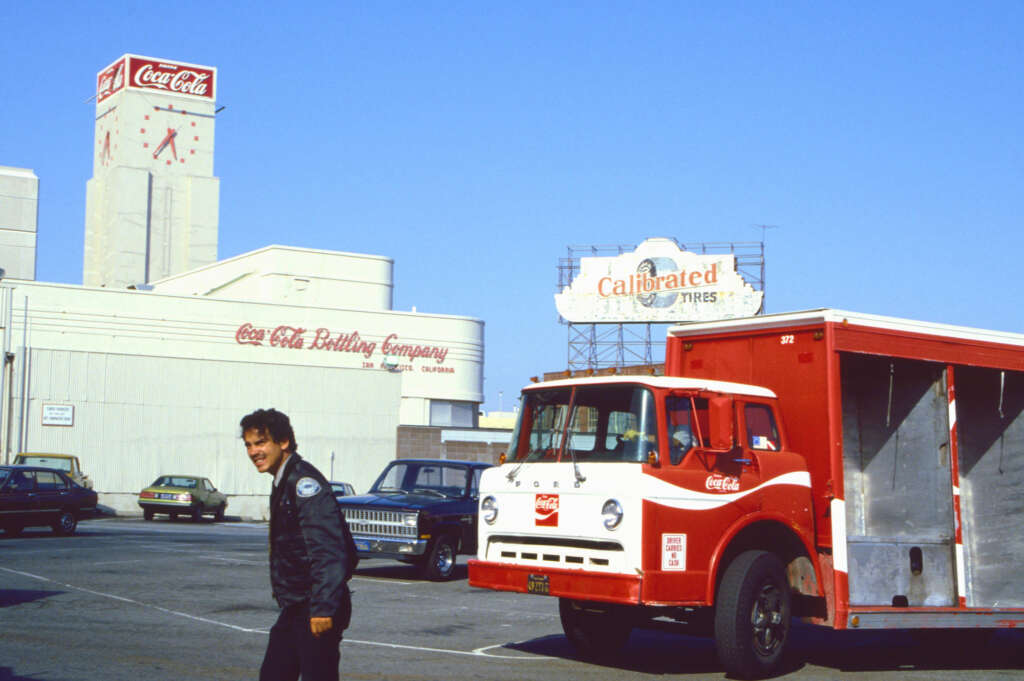 A man walks near a vintage Coca-Cola delivery truck in a parking lot. Buildings with Coca-Cola and Califoam Tires signage appear in the background under a clear blue sky. Several cars are parked nearby.