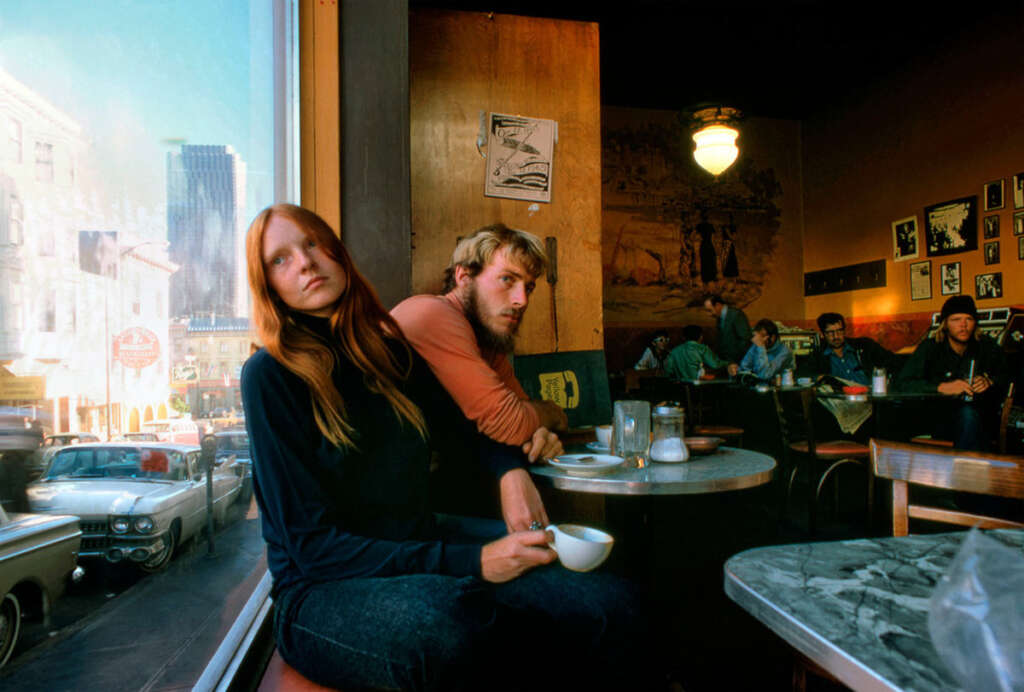 A woman with long red hair and a man with blonde hair sit at a marble table in a dimly lit café, holding cups. Through the large window, city buildings and parked cars are visible. Other patrons are seated in the background.