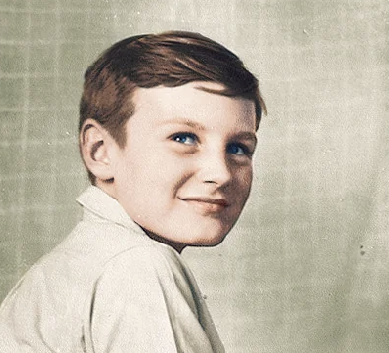 A young boy with short brown hair and blue eyes gazes slightly to the right. He is wearing a light-colored shirt. The background is a plain, muted color, creating a vintage or nostalgic feel to the portrait.