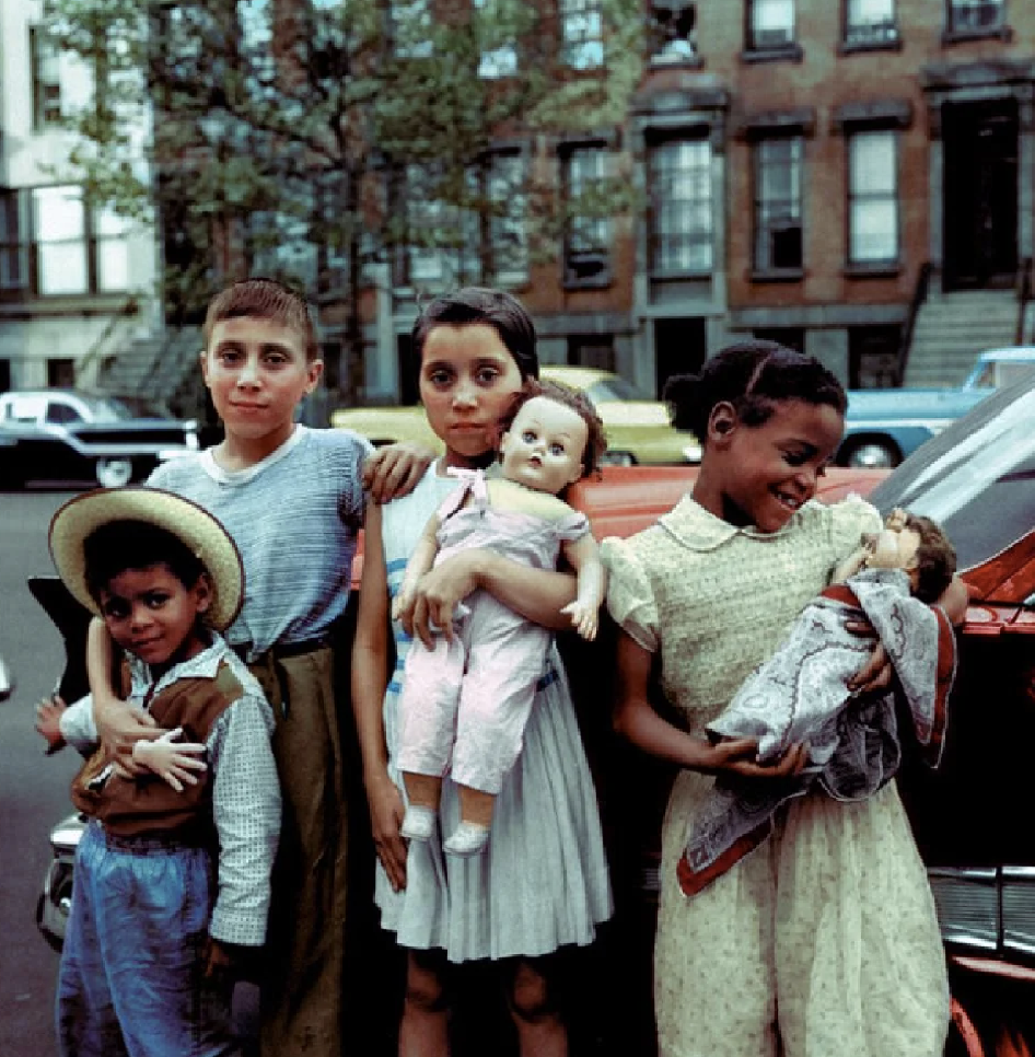 Four children stand in front of a parked car on a city street. Two of them hold dolls. They are dressed in mid-20th-century clothing, with a backdrop of brownstone buildings and parked vintage cars. The group looks directly at the camera.