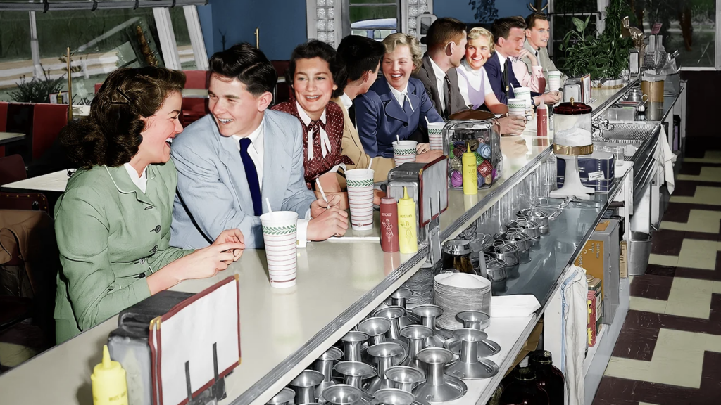 A retro diner scene with young people sitting at a long counter, chatting and smiling. The counter is lined with condiments, cups, and dessert displays. The floor has a checkered pattern, and there's a window view in the background.