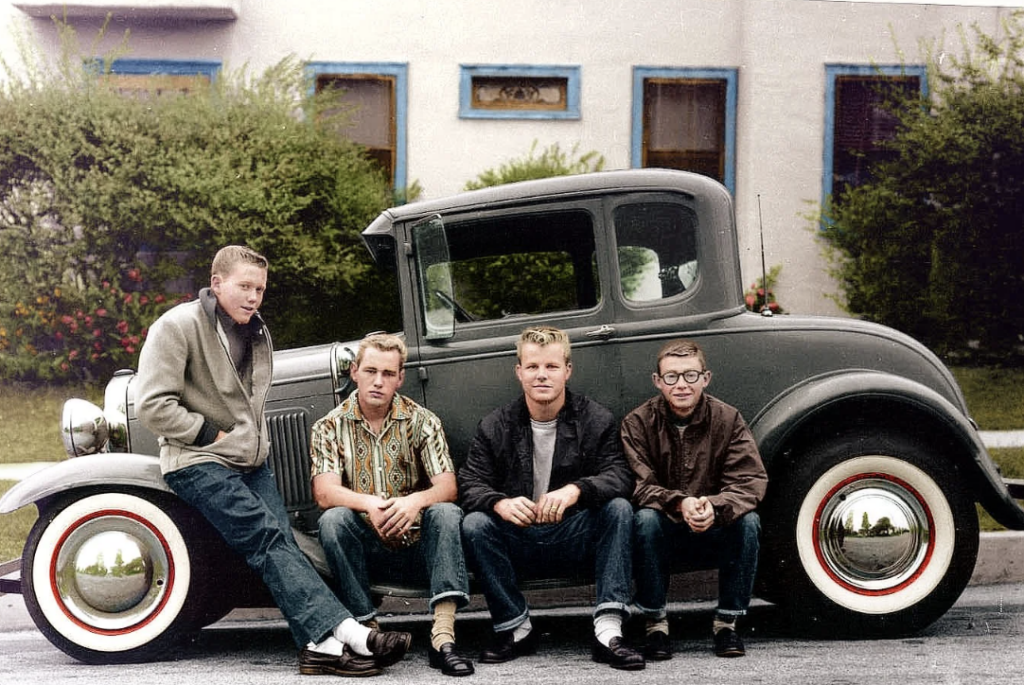 Four young men sitting in front of a vintage car on a residential street. They are dressed in casual 1950s-style clothing with jeans and jackets. The car has white wall tires, and a house with bushes and windows is visible in the background.