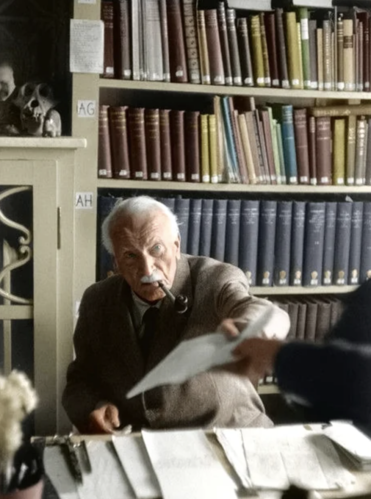 An older person with a mustache is sitting at a desk, holding a pipe in their mouth. They're reaching for a document handed to them by someone off-camera. Shelves filled with books are in the background.